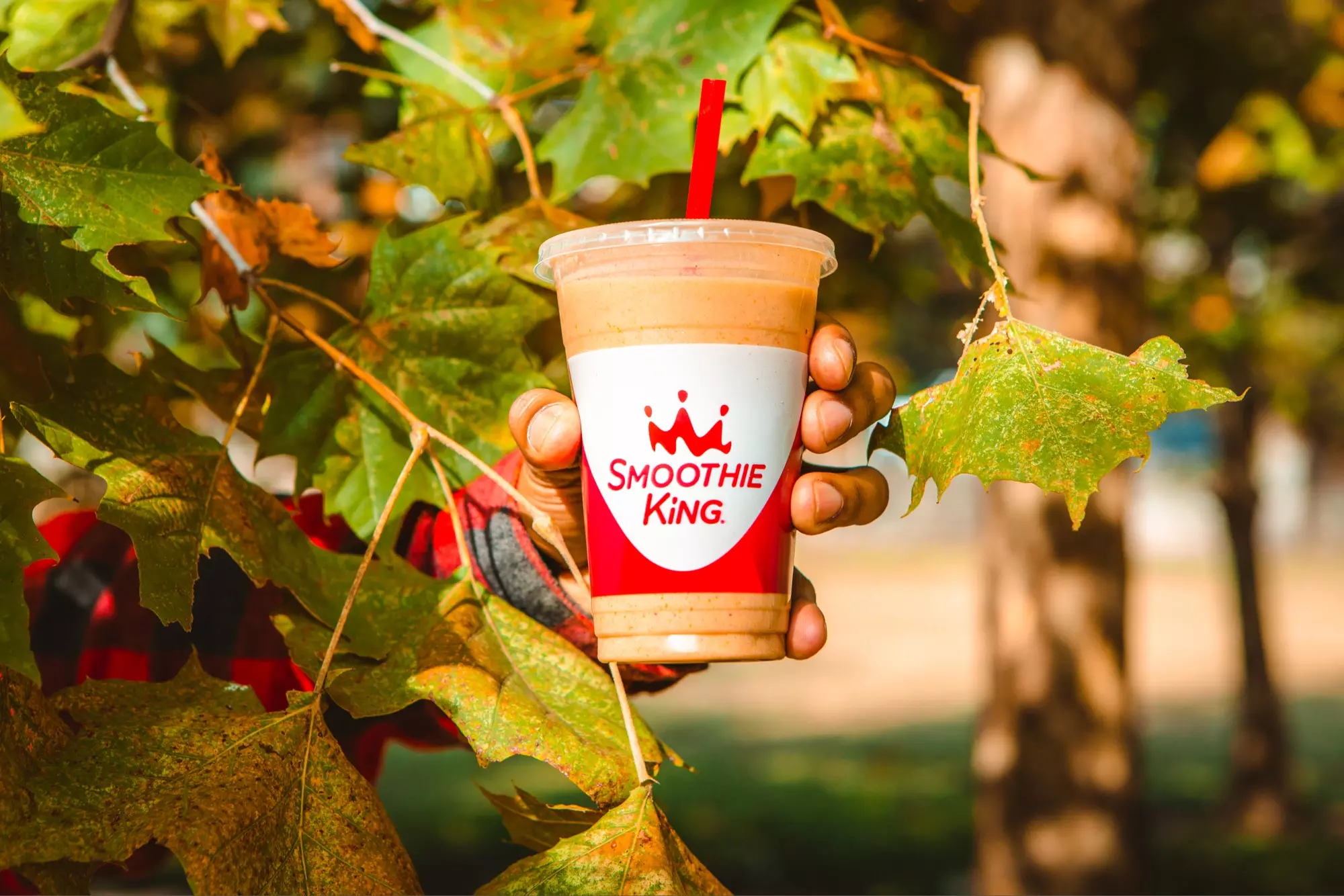 A person holding a Smoothie King pumpkin smoothie in front of leaves on a tree in the fall